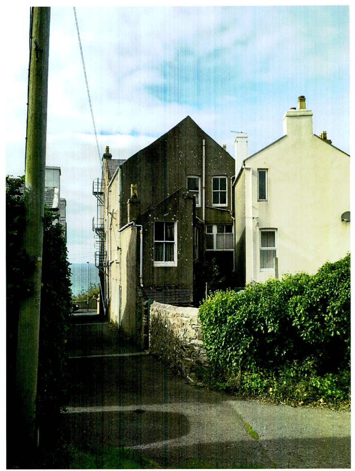 A street-level photograph showing a dark two-story building next to a white house, with a stone wall and sea view in the background.
