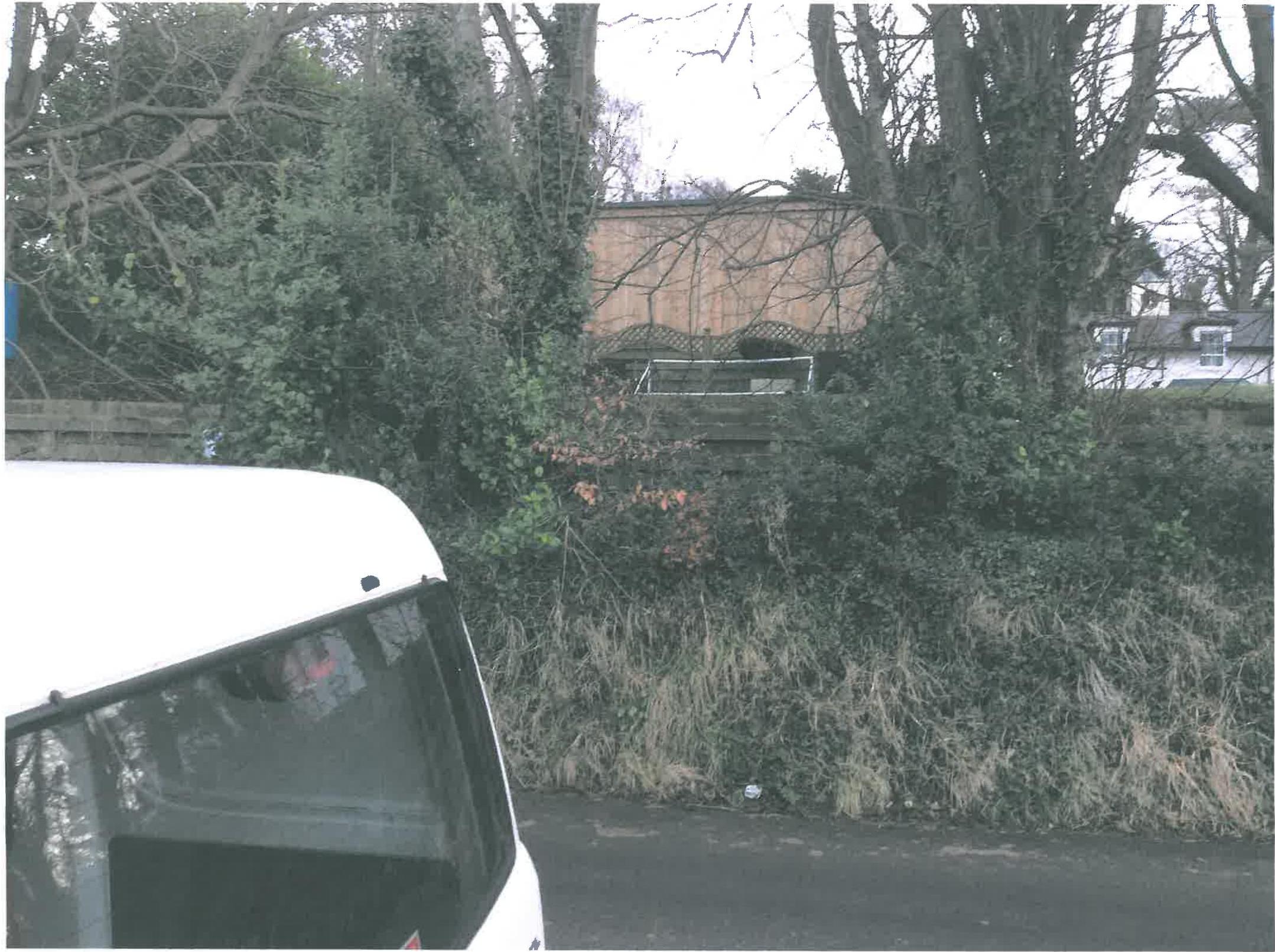 A photograph taken from a street level showing a wooden outbuilding or summerhouse partially obscured by trees and a fence. A white vehicle is visible in the foreground.