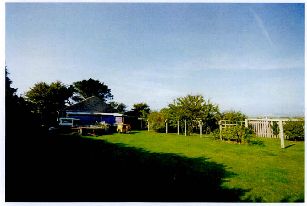 A photograph showing a grassy rear garden area with a building on the left and a wooden trellis fence on the right.