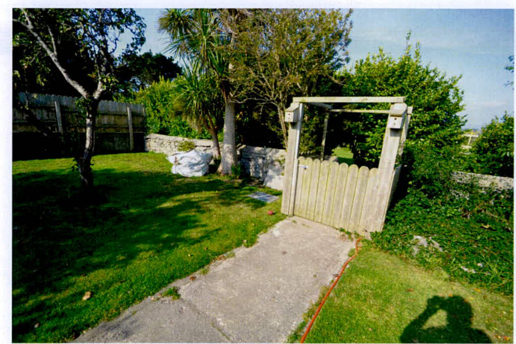 A photograph showing a grassy rear garden area with a wooden gate, concrete path, and surrounding trees and stone walls.