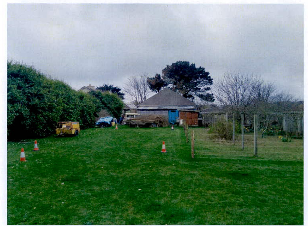 A photograph showing a grassy rear garden area with traffic cones marking a boundary, leading towards a barn-like outbuilding with a blue door and a large hedge on the left.