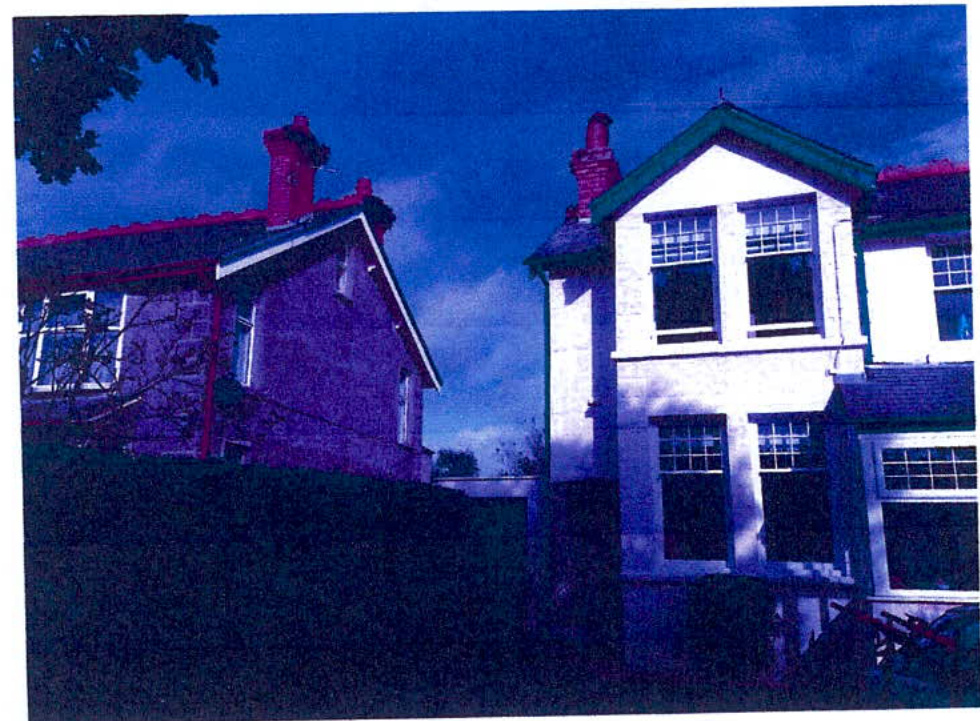 A photograph showing the exterior of a two-story white house with green trim and an adjacent building, likely the existing property for the extension application.