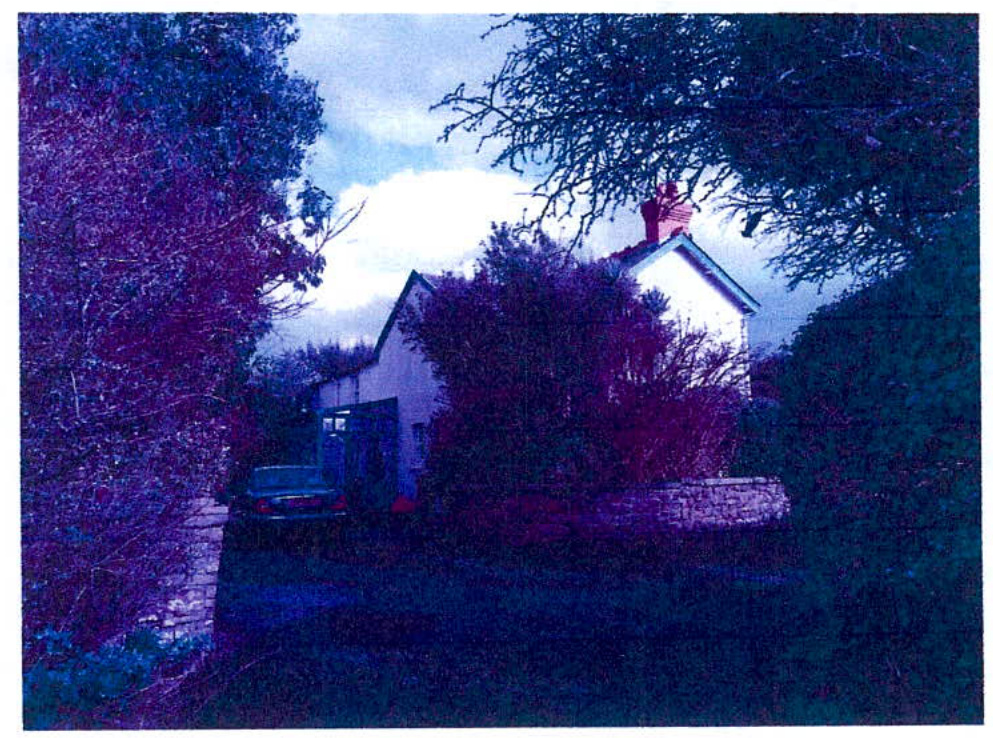 A grainy, blue-tinted photograph showing the exterior of a white residential property with a stone wall and surrounding vegetation.