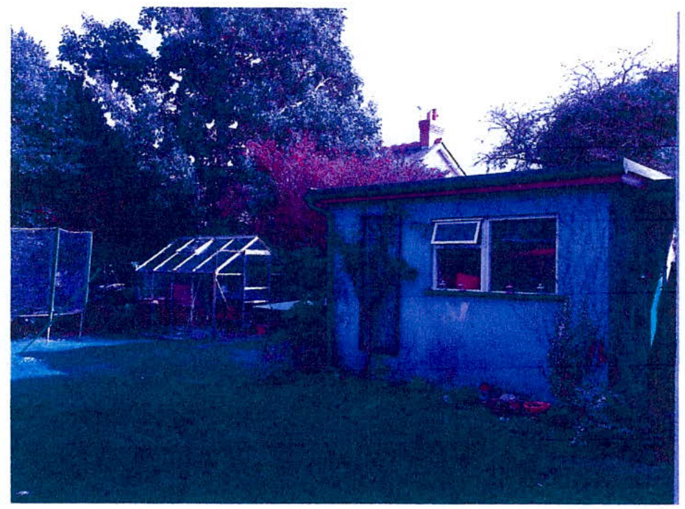 A low-quality photograph of a rear garden featuring a small outbuilding, a greenhouse, and a trampoline with trees in the background.