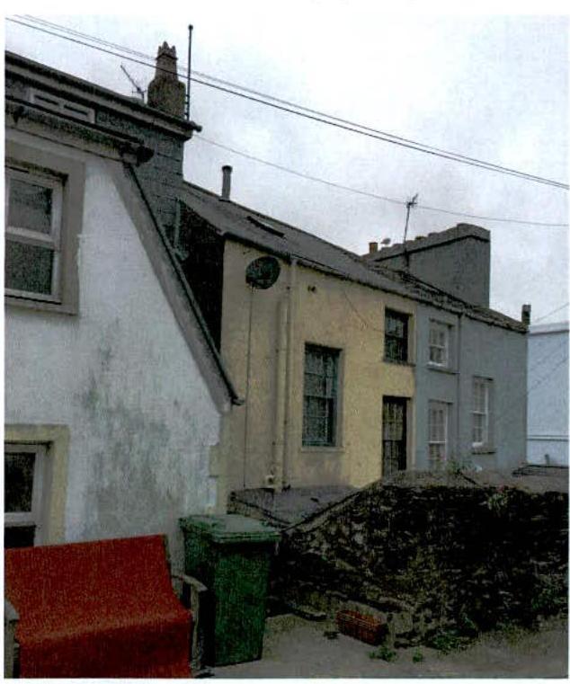 A photograph showing a row of terraced buildings with a stone wall and waste bins in the foreground.