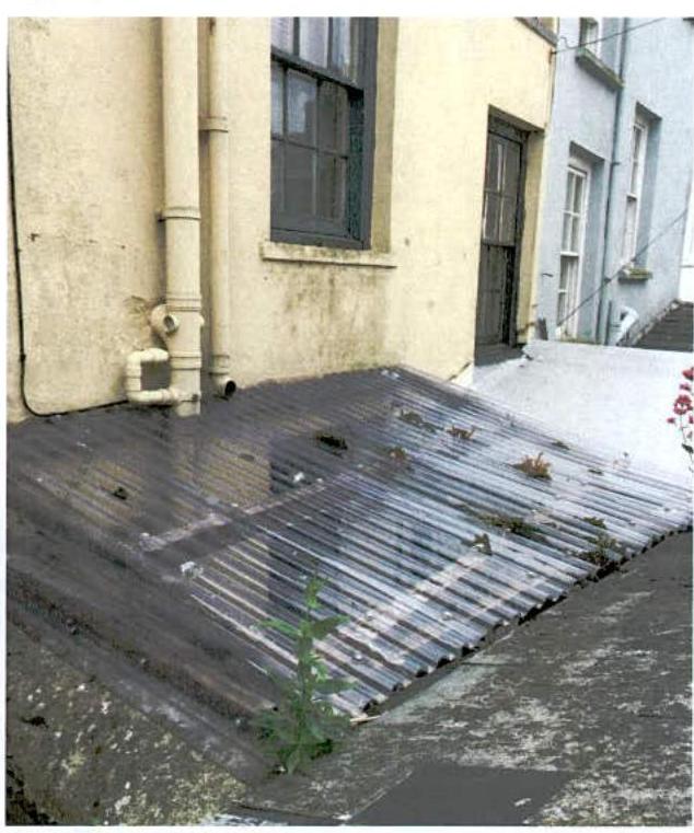 A photograph showing a dilapidated corrugated roof structure, likely a garage or lean-to, attached to the side of a building with weeds growing through it.