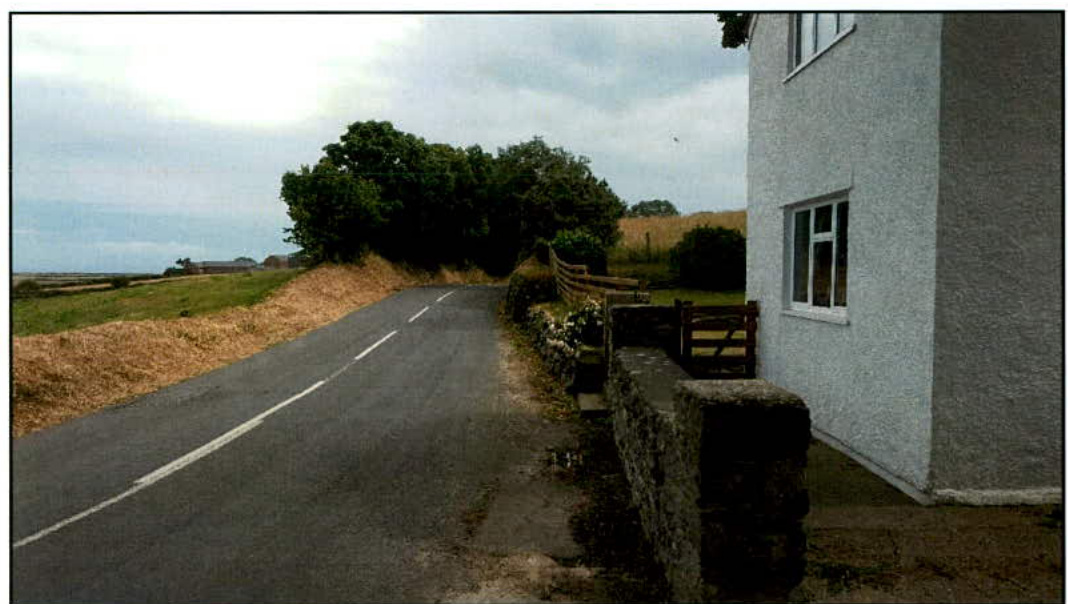 A photograph showing a paved rural road curving past a white building with a stone wall and wooden gate on the right side.