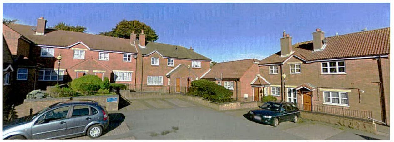 A street-level photograph showing a row of red brick terraced or semi-detached houses with parked cars and landscaping.