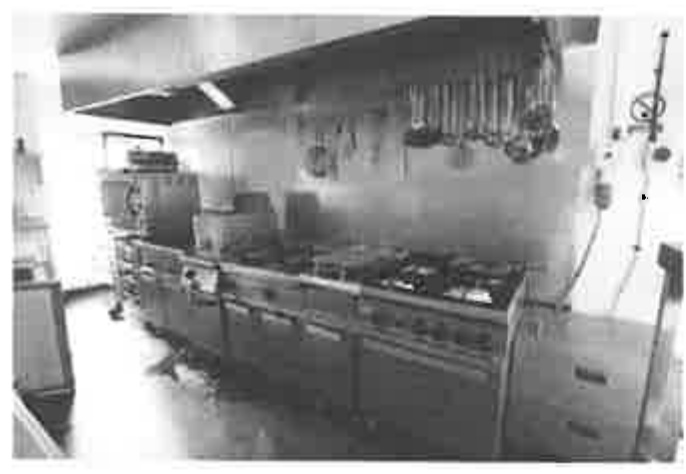 Black and white photograph showing the interior of a commercial kitchen with a large stainless steel range and extractor hood.