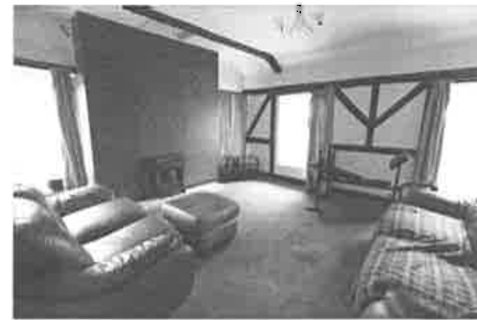 A grainy black and white photograph showing the interior of a room with exposed timber framing, sofas, and large windows.