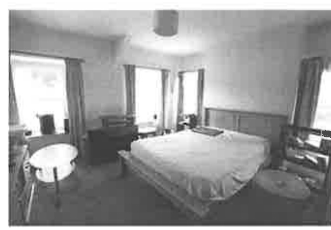 A grainy black and white photograph showing the interior of a bedroom with a double bed, windows with curtains, and furniture.