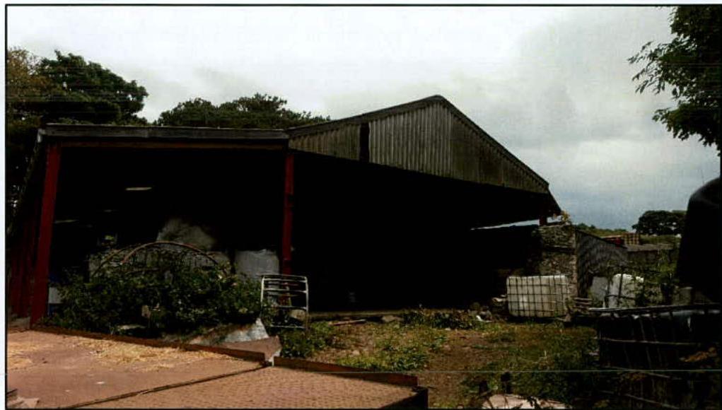 A photograph of a large, open-sided agricultural building or shed with a corrugated roof, situated in a rural area.