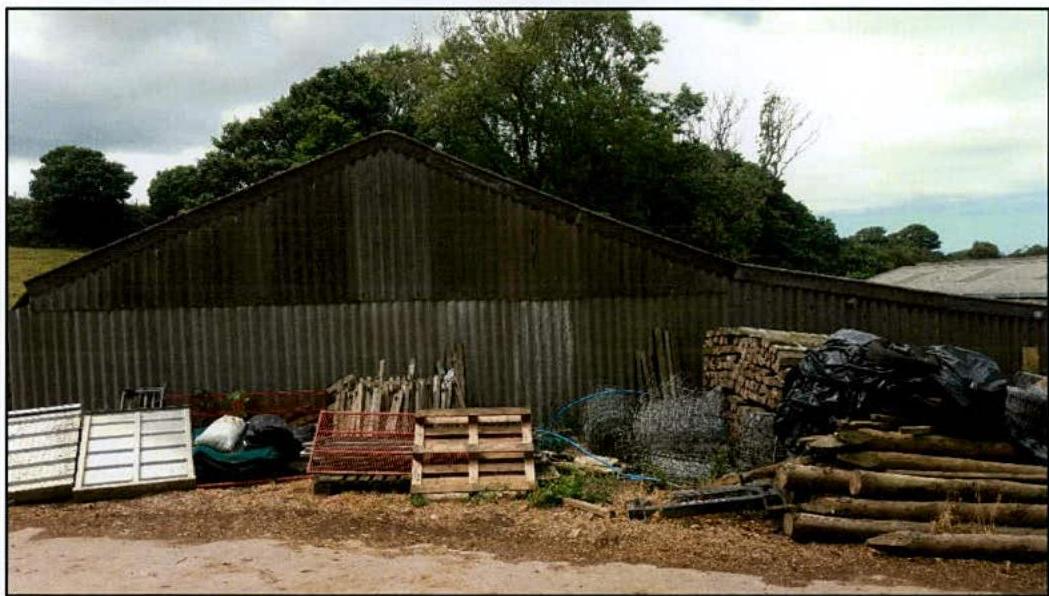 A photograph showing a large corrugated metal agricultural building or barn situated in a rural setting with timber and materials stacked in the foreground.