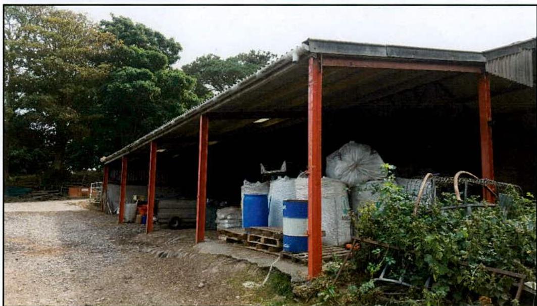 A photograph showing an existing open-sided agricultural building with red steel supports, containing stored sacks and barrels, situated in a rural setting.