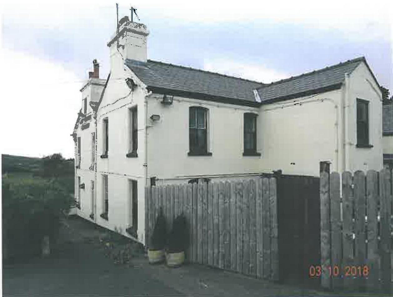 A photograph showing the exterior of a white, two-story building with a slate roof and chimney stacks. A wooden fence runs along the front of the property.