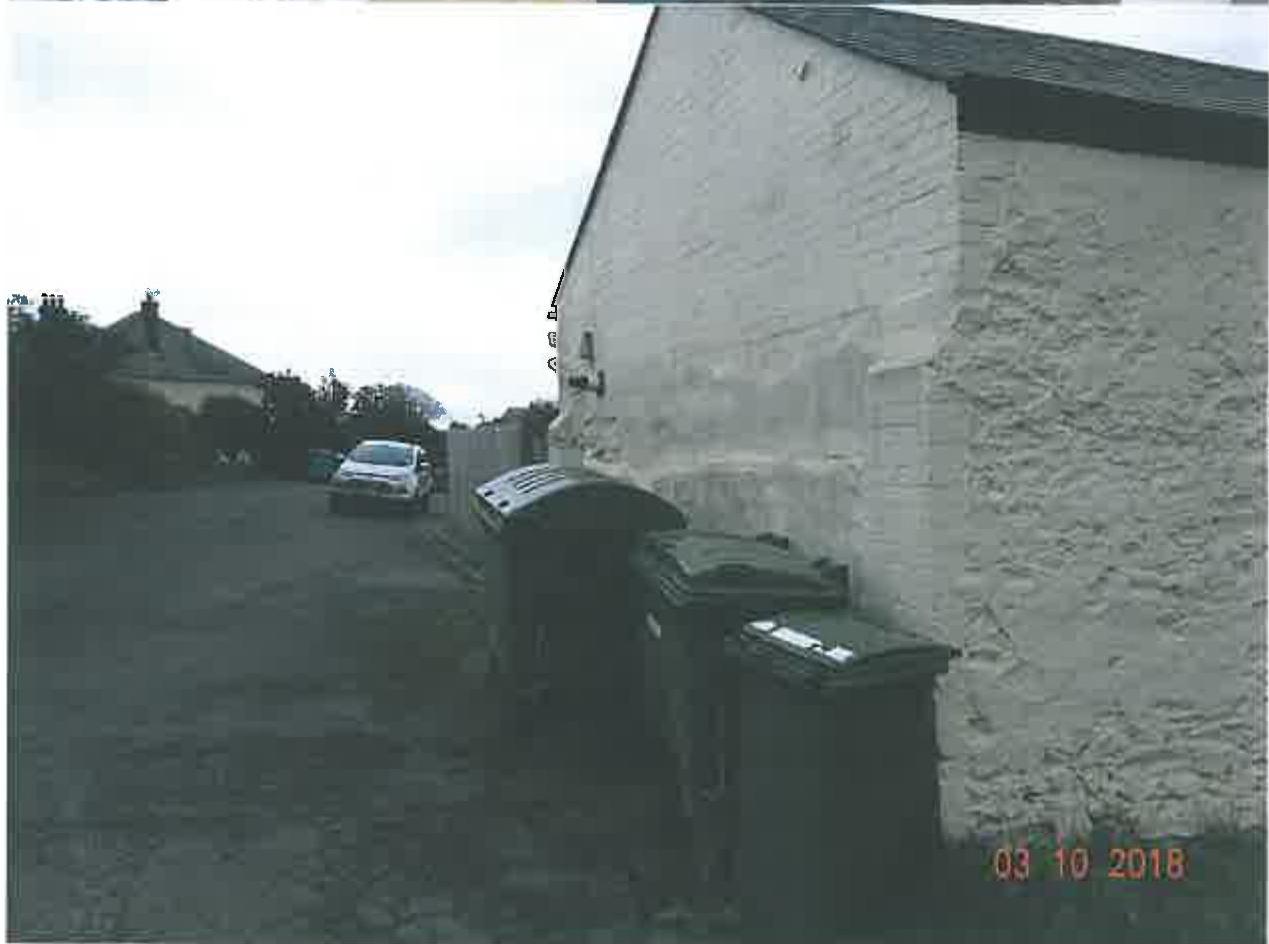 A photograph showing the side elevation of a white-washed building with several large waste bins lined up against the wall and a car parked on the driveway in the background.