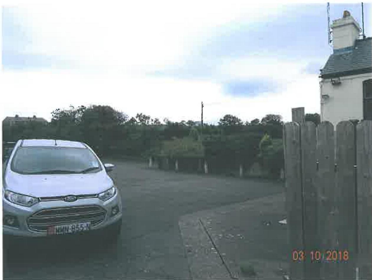 A photograph showing a white car parked on a paved forecourt next to a wooden fence and the corner of a white building.