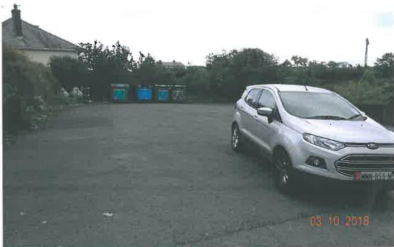 A photograph showing a paved car park area with a silver car parked on the right and several large waste bins in the background.