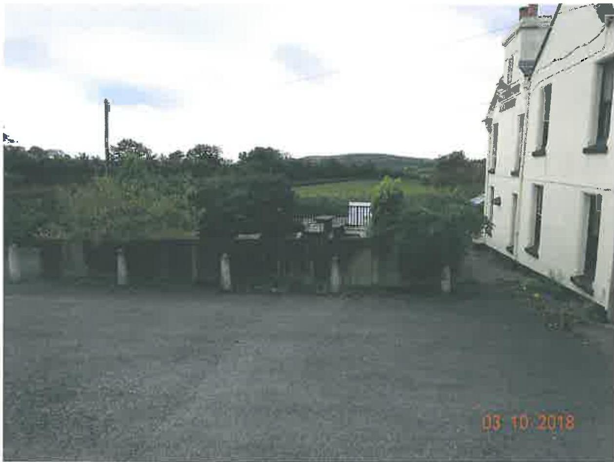A photograph showing the side elevation of a white two-story building with a large paved area in the foreground and rural scenery in the background.
