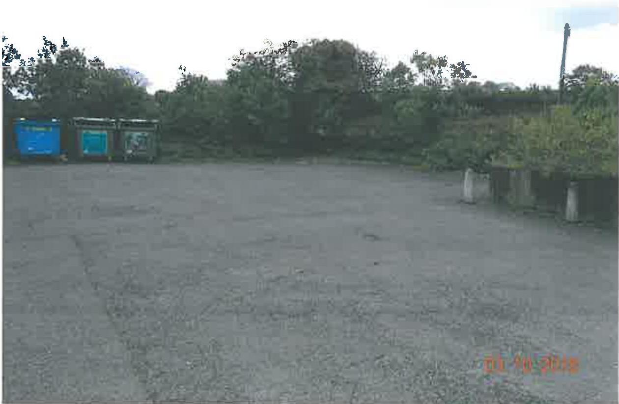 A photograph showing a paved parking area or forecourt with waste bins on the left and trees in the background.