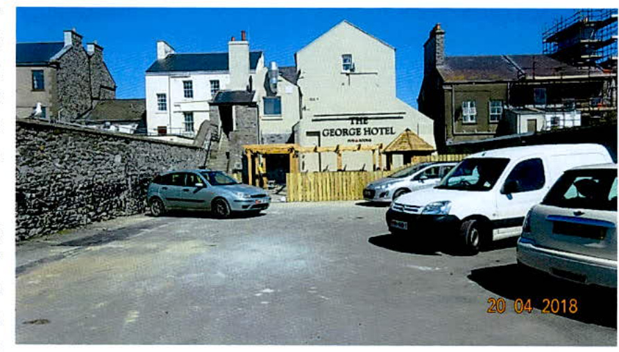 A photograph showing a parking area with cars, a stone wall, and a new wooden fence in front of a building labeled 'The George Hotel'.