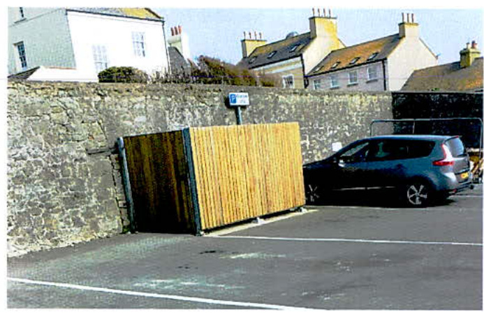 A photograph showing a wooden fence panel leaning against an existing stone wall in a paved area, with a car parked nearby and residential buildings in the background.
