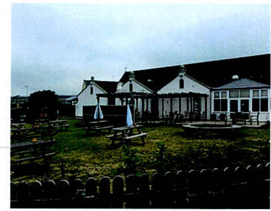 A photograph showing the rear exterior of a white commercial building, likely a pub, with an outdoor patio area featuring picnic tables and a wooden fence in the foreground.
