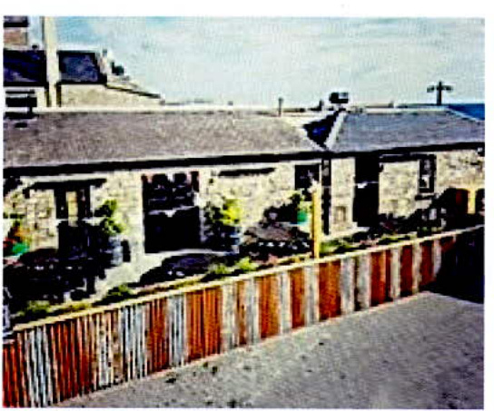 A low-resolution photograph showing the rear of a stone building with a wooden fence installed along the boundary.