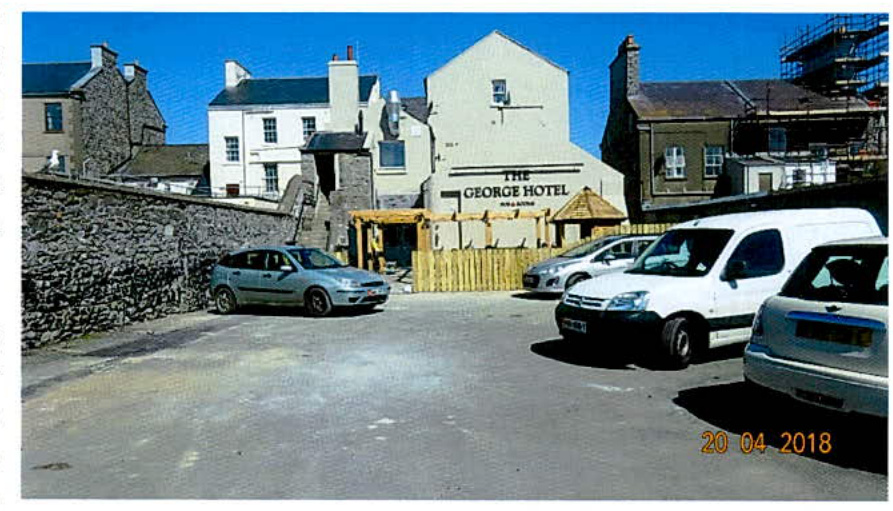 A street-level photograph showing a paved parking area with several cars and a new wooden fence in the mid-ground. The background features a white building identified as 'The George Hotel' and adjacent stone structures.