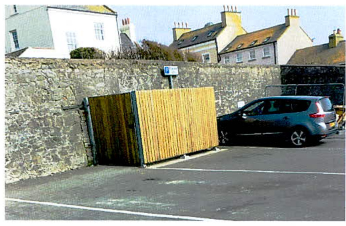 A photograph showing a newly installed wooden fence panel next to a stone wall in a paved area, with a car parked nearby and residential buildings in the background.