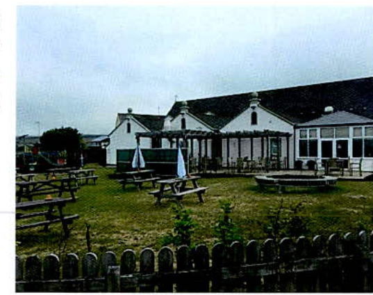 A photograph showing the rear garden area of a commercial building, likely a pub, featuring picnic tables, a wooden fence in the foreground, and a conservatory extension.
