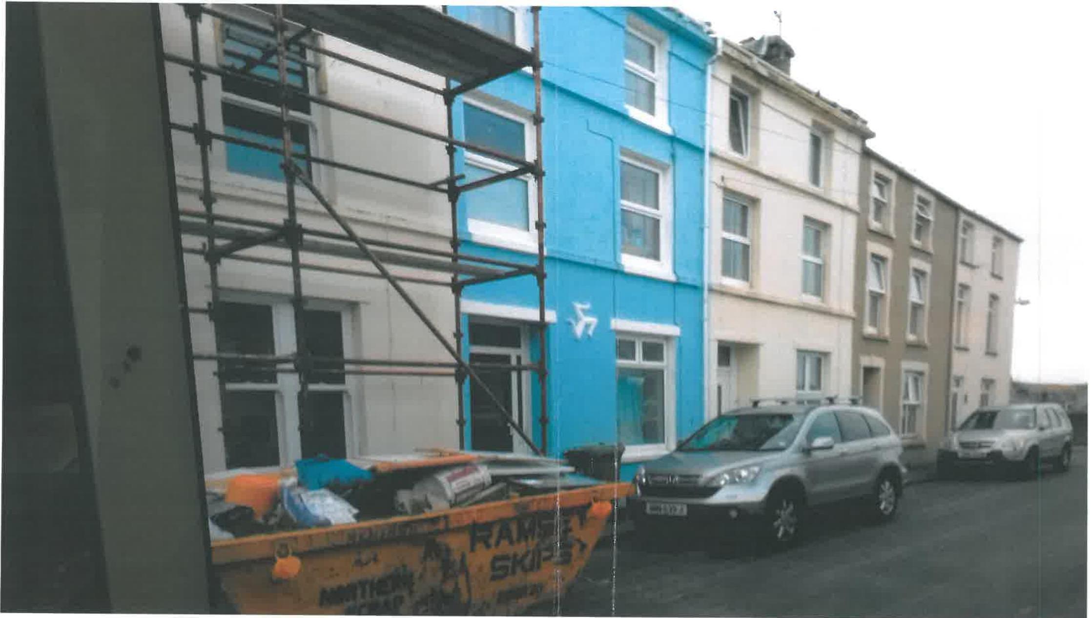 A street-level photograph showing a row of terraced houses, with the leftmost building covered in scaffolding and a skip in the foreground.