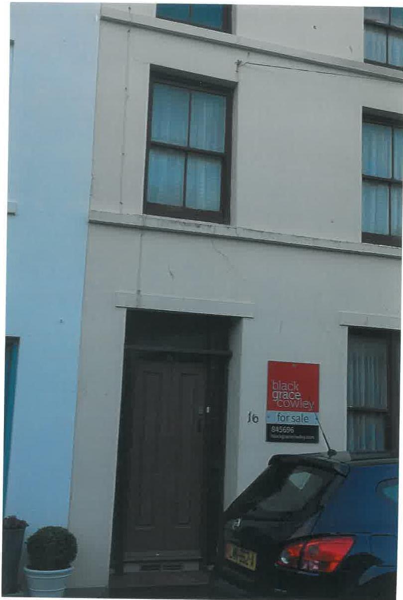 A photograph showing the front elevation of a white terraced house with a dark door and sash windows. A 'for sale' sign is attached to the wall next to the entrance, and a blue car is parked in the foreground.