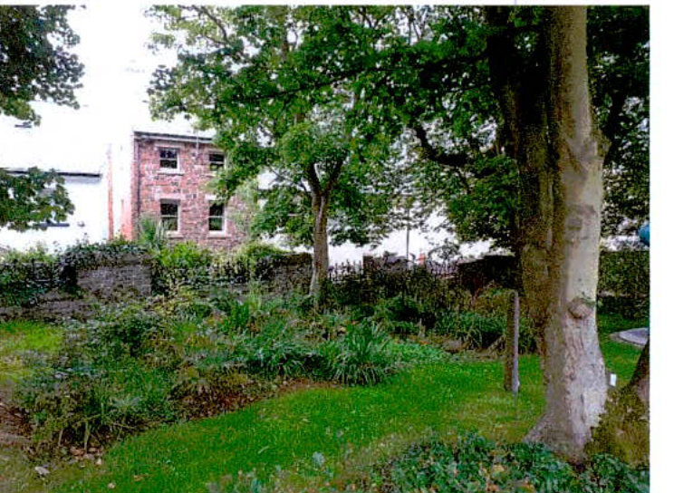 A photograph showing a grassy garden area with trees and a stone wall, with a brick building visible in the background.