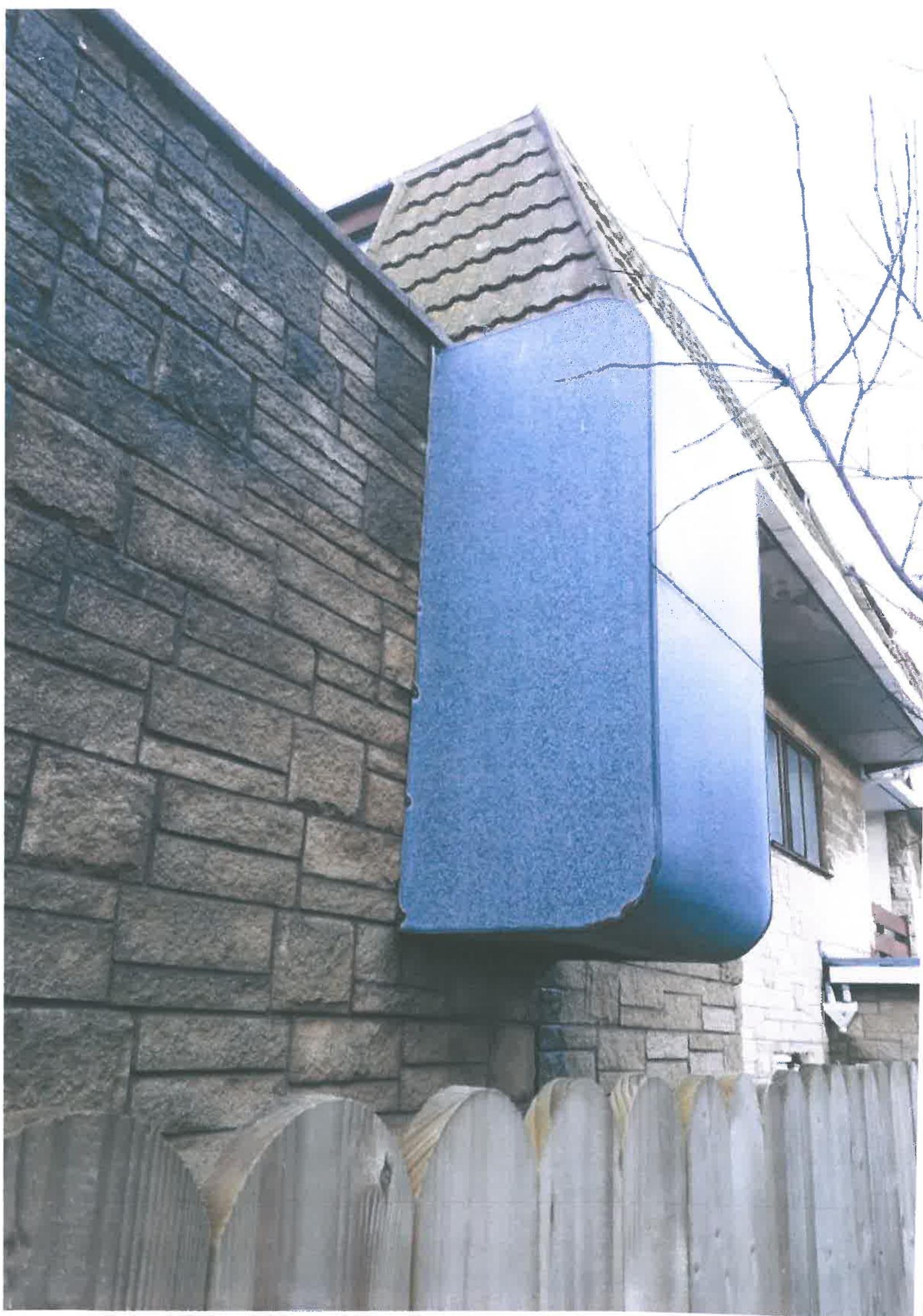 A low-angle photograph showing the exterior of a building with a stone wall, a large blue protruding structure, and a wooden fence in the foreground.