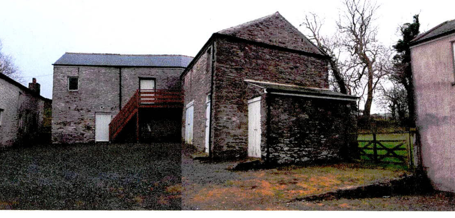 A photograph showing a cluster of stone buildings, including a two-story structure with an external wooden staircase and an adjacent barn-like outbuilding with white doors.