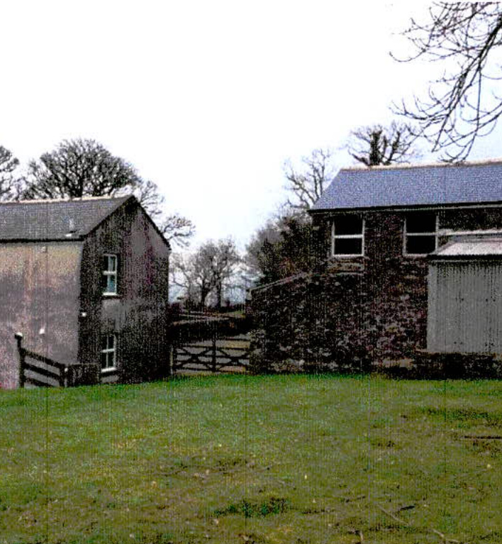 A grainy exterior photograph showing a grey outbuilding on the left and a stone house on the right, separated by a wooden gate in a rural setting.