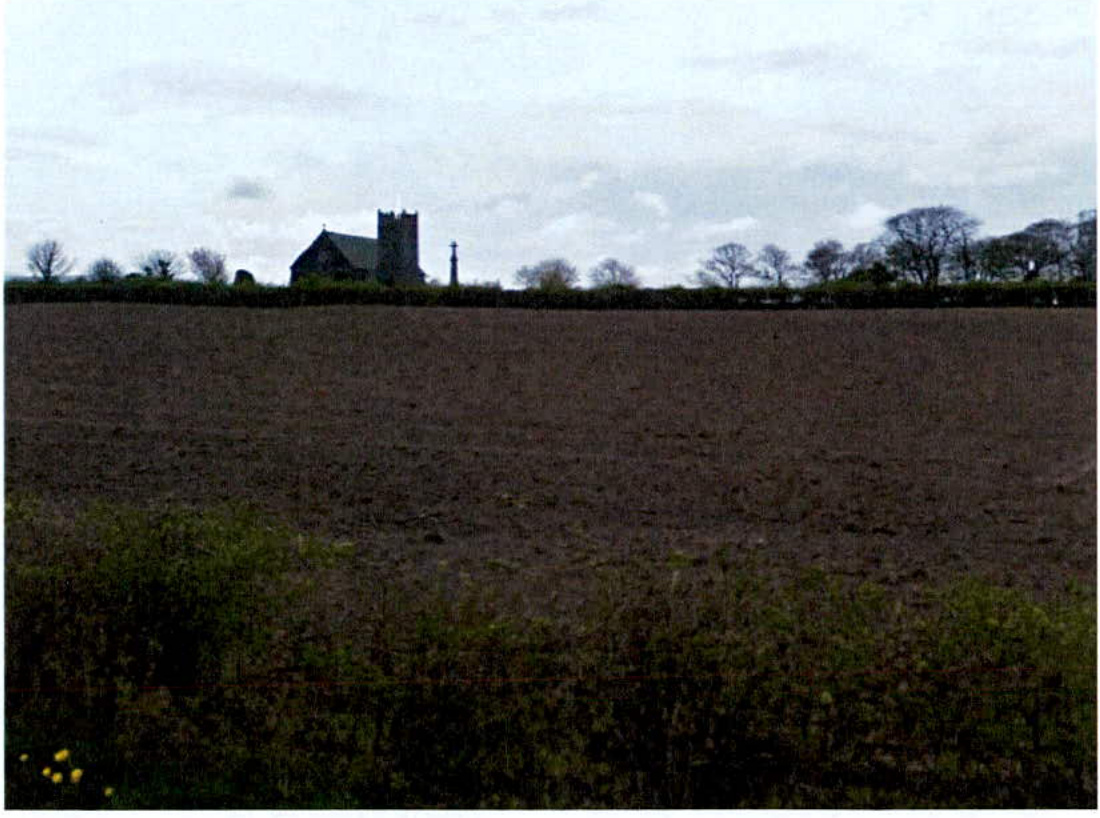 A photograph showing a rural landscape with a ploughed field in the foreground and a church with a tower visible on the horizon behind a hedge.