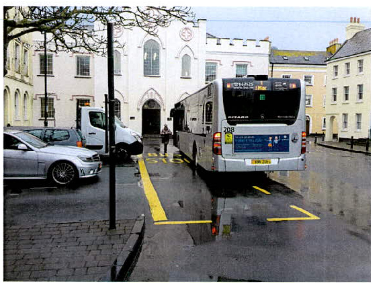 A street-level photograph showing a white bus stopped at a designated bus bay marked with yellow road markings. The scene includes parked cars on the left and white buildings with arched windows in the background.
