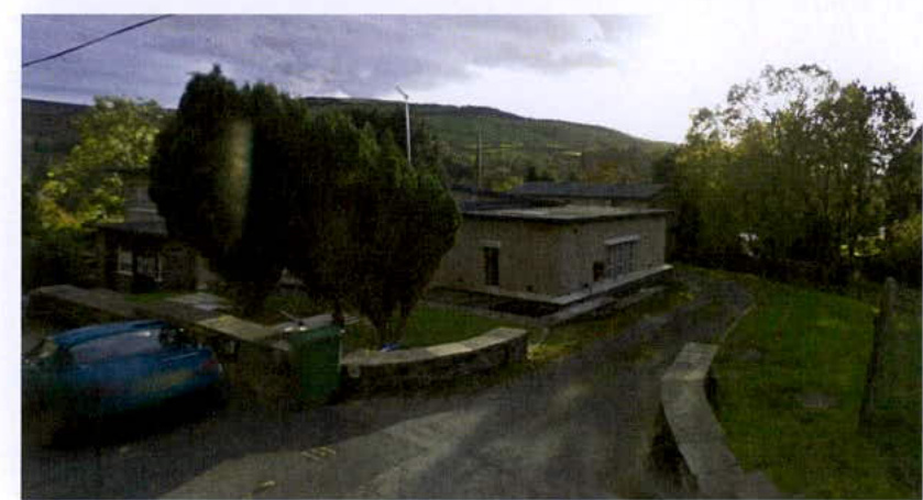 A grainy exterior photograph showing a single-story building with a flat roof, situated in a rural setting with a driveway and a parked blue car.