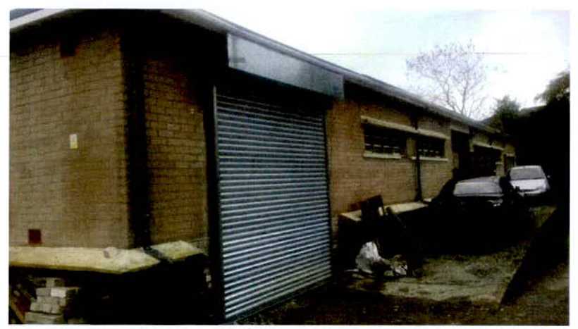 A photograph showing the exterior of a single-story brick building featuring a large metal roller shutter door and parked vehicles.
