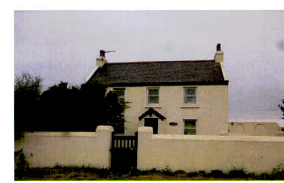 A photograph of a white, two-story detached house with a dark tiled roof, situated behind a white boundary wall with a black gate.