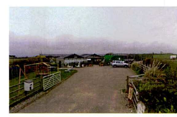 A grainy photograph showing a paved driveway leading towards existing buildings in a rural setting.