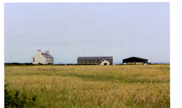A photograph showing a rural field with a white detached house and two agricultural buildings in the distance.