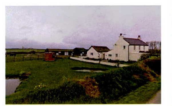 A photograph showing a rural property with white buildings and green fields, likely depicting the existing site conditions for the proposed development.