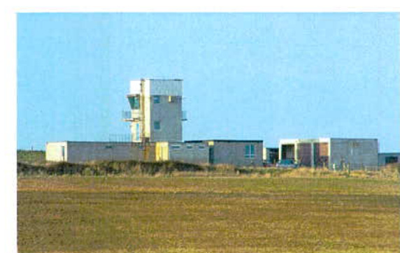 A grainy photograph showing a building complex with a prominent tower structure, likely an airfield control tower, situated in a grassy field under a blue sky. This image serves as a context view for the proposed deta...