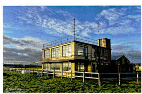 A photograph of a two-story building resembling an old airfield control tower, situated in a grassy field with a wooden fence.