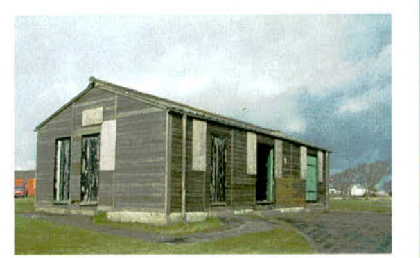 A photograph showing a long, single-story wooden building, likely a temporary structure or outbuilding, situated in a grassy rural field.