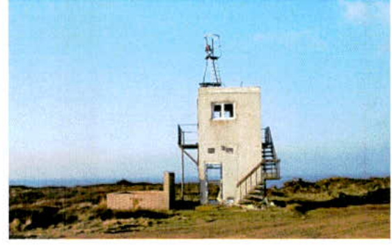 A photograph showing a small concrete tower-like structure with an external staircase situated in a grassy coastal landscape with the sea visible in the background.