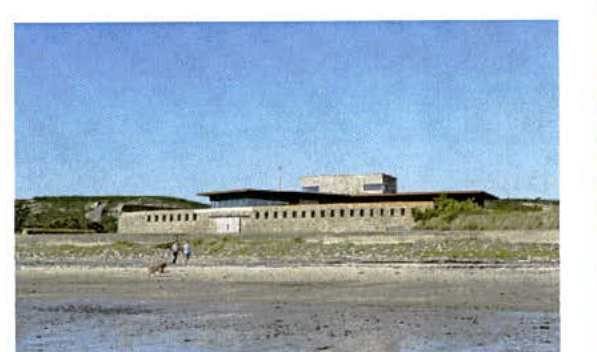 A photograph showing a long, low concrete building situated on a coastal dune or beach area with people walking in the foreground.
